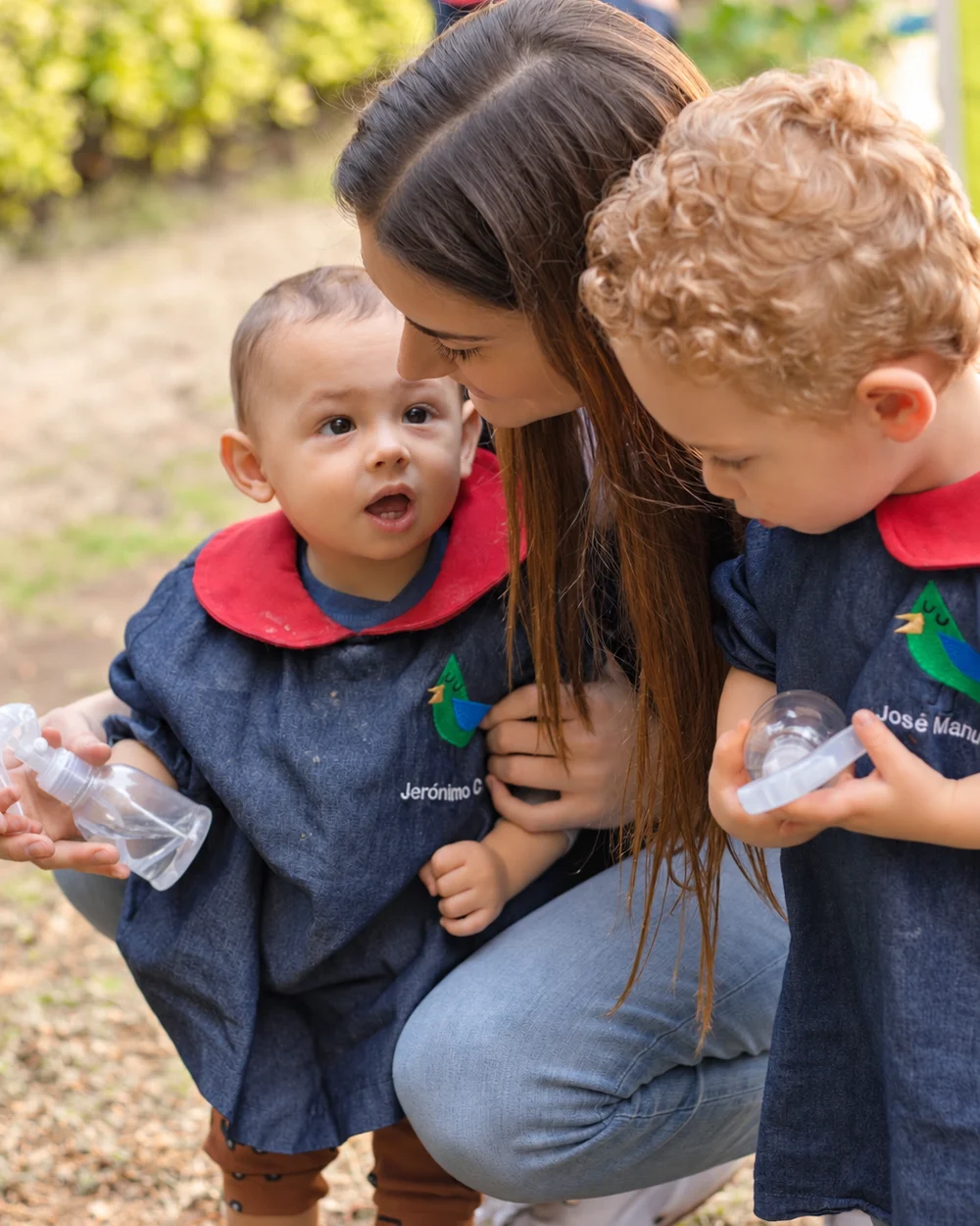 Maestra de Pirulí hincada en el jardín platicando con dos alumnos pequeños que sostienen sus botellas de agua
